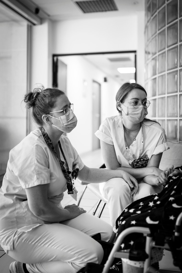 Photographie en noir et blanc de Sophie, infirmière de soins palliatifs, et Laura, aide-soignante en soins palliatifs. Elles portent un masque et sont accroupies devant une patiente, hors du cadre de la photo. Laura lui tient la main.