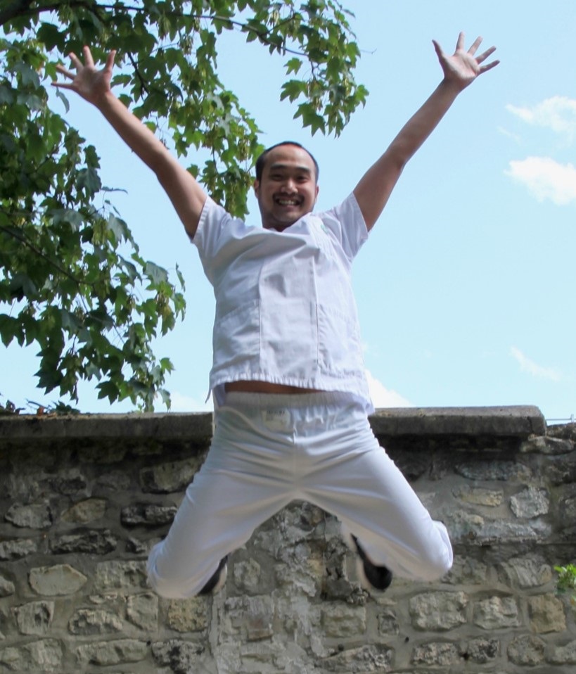 Photographie d'Alexandre, kinésithérapeute. Il est dehors, en blouse de soignant, et fait un grand saut avec les mains vers le ciel, en souriant