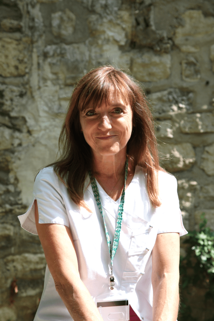 Photographie de Corinne, socio-esthéticienne. Elle est dehors, au soleil, en blouse de soignante, et sourit