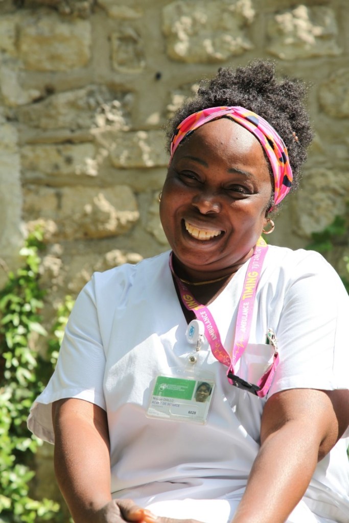 Photographie de Nogaye, agente de service hospitalier. Elle est dehors, au soleil, en blouse de soignante, et sourit