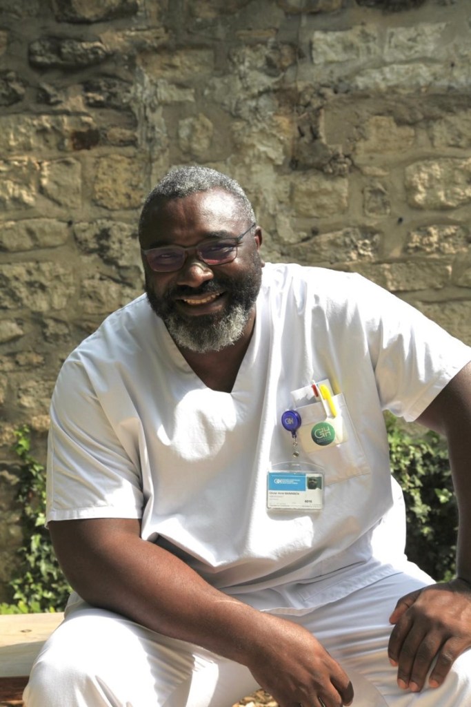 Photographie d'Olivier, aide-soignant. Il est dehors, au soleil, en blouse de soignant, et sourit