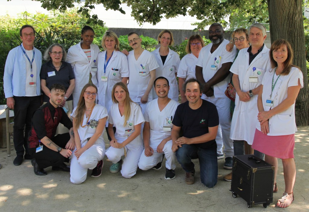 Photographie de 16 membres de l'équipe de soins palliatifs. Ils sont regroupés dehors, sous un arbre. Certains portent une blouse de soignant, d'autres sont en tenue civile. Ils regardent l'objectif et sourient